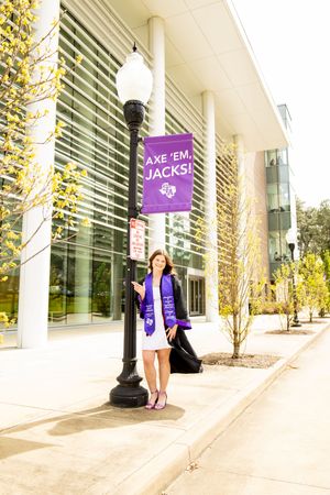 College graduate in white dress with purple stole poses by Axe Em Jacks banner on campus lamppost.