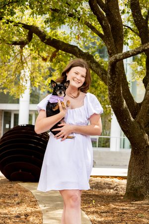 Smiling young woman in white dress holding a small black Chihuahua dog outdoors under a tree.