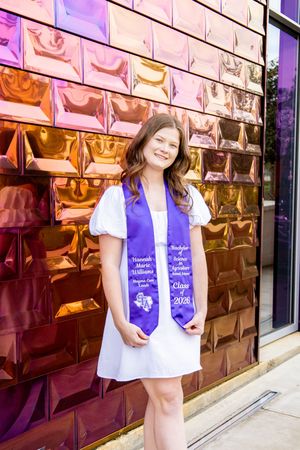 Young woman in white dress wearing purple graduation stole posing in front of colorful metallic wall.