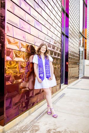 Young woman in white dress and purple graduation stole posing against reflective metallic wall.