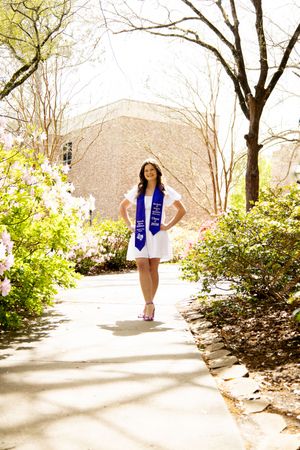 Smiling graduate in white dress and blue stole posing on sunny campus pathway with blooming flowers.