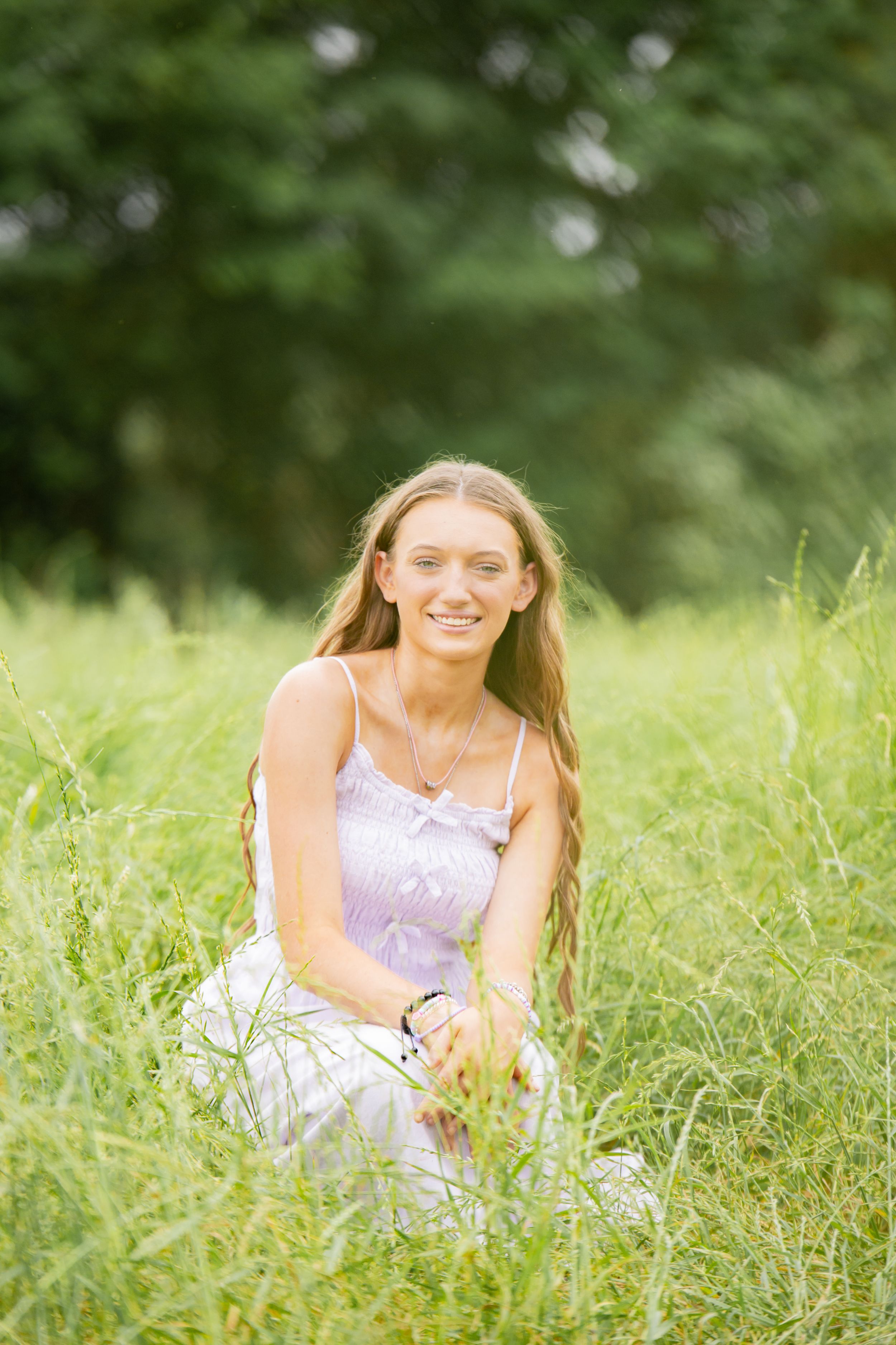 Smiling young woman with long hair sitting in lush green grass outdoors on a sunny day.