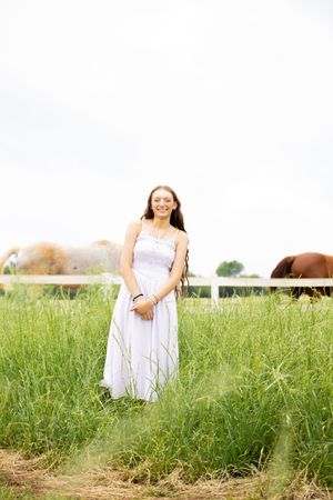 Young woman in white sundress smiling in green field with horses grazing behind white fence.