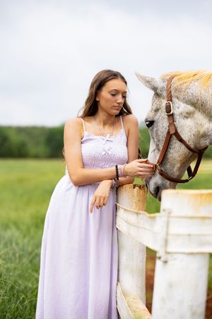 Young woman in lavender maxi dress gently interacts with a gray horse at a white fence in a green field.