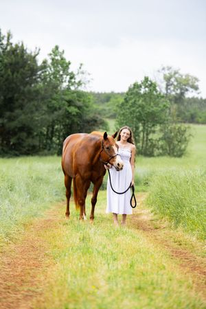 Young woman in white dress leads a chestnut horse along a rural dirt path surrounded by lush green fields.