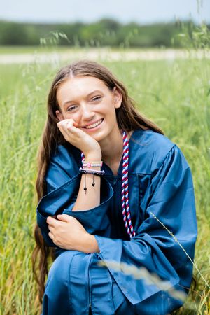 Smiling young woman in blue graduation gown sitting in green field, wearing honor cords and bracelets.