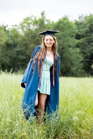 Happy graduate in blue cap and gown standing in tall green grass outdoors.
