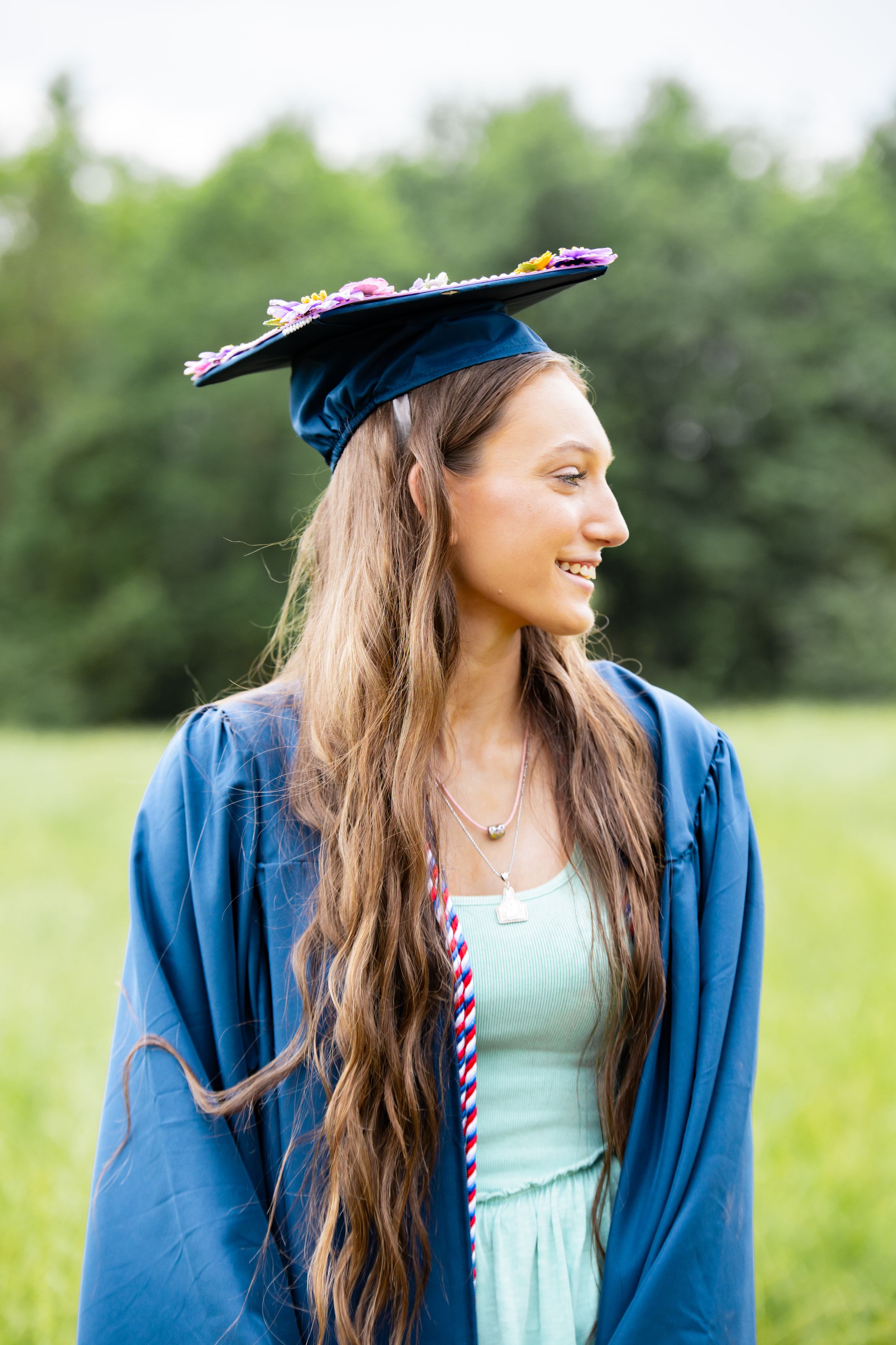 Smiling young woman in blue graduation cap and gown standing outdoors in a green field.