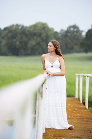 Young woman in white maxi dress stands on a wooden bridge overlooking a serene green meadow landscape.