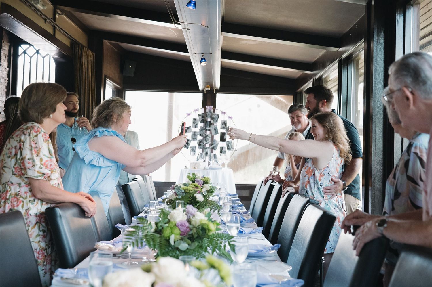 People raise champagne glasses in a toast around an elegant table setting with white floral centerpieces on a boat.