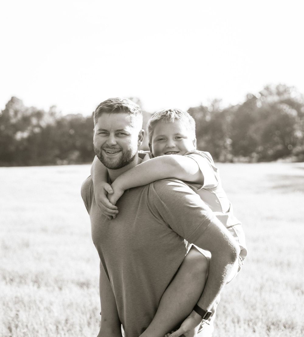 A heartwarming piggyback ride moment captured in black and white in a sunny outdoor field.