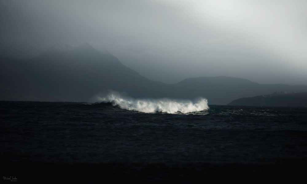 Dramatic ocean waves crash beneath dark stormy mountains covered in moody mist and clouds.