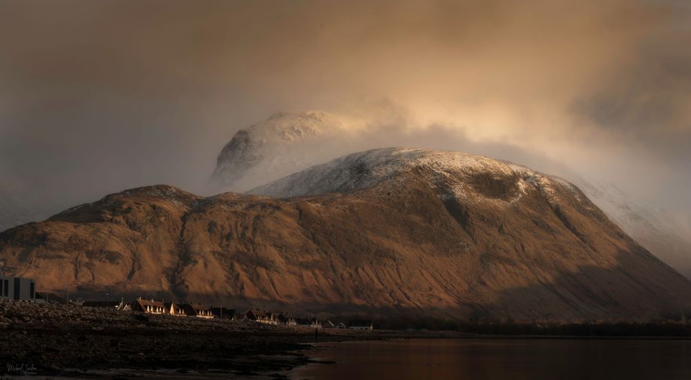 Dramatic mountain peak shrouded in misty clouds during golden hour with rocky terrain in the foreground.