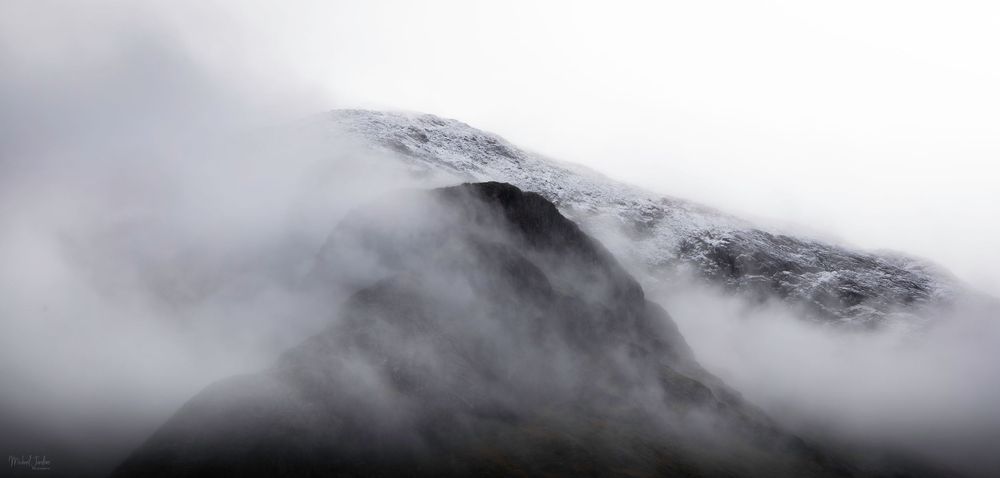 Misty clouds swirl around a snow-capped mountain peak in dramatic black and white landscape photography.