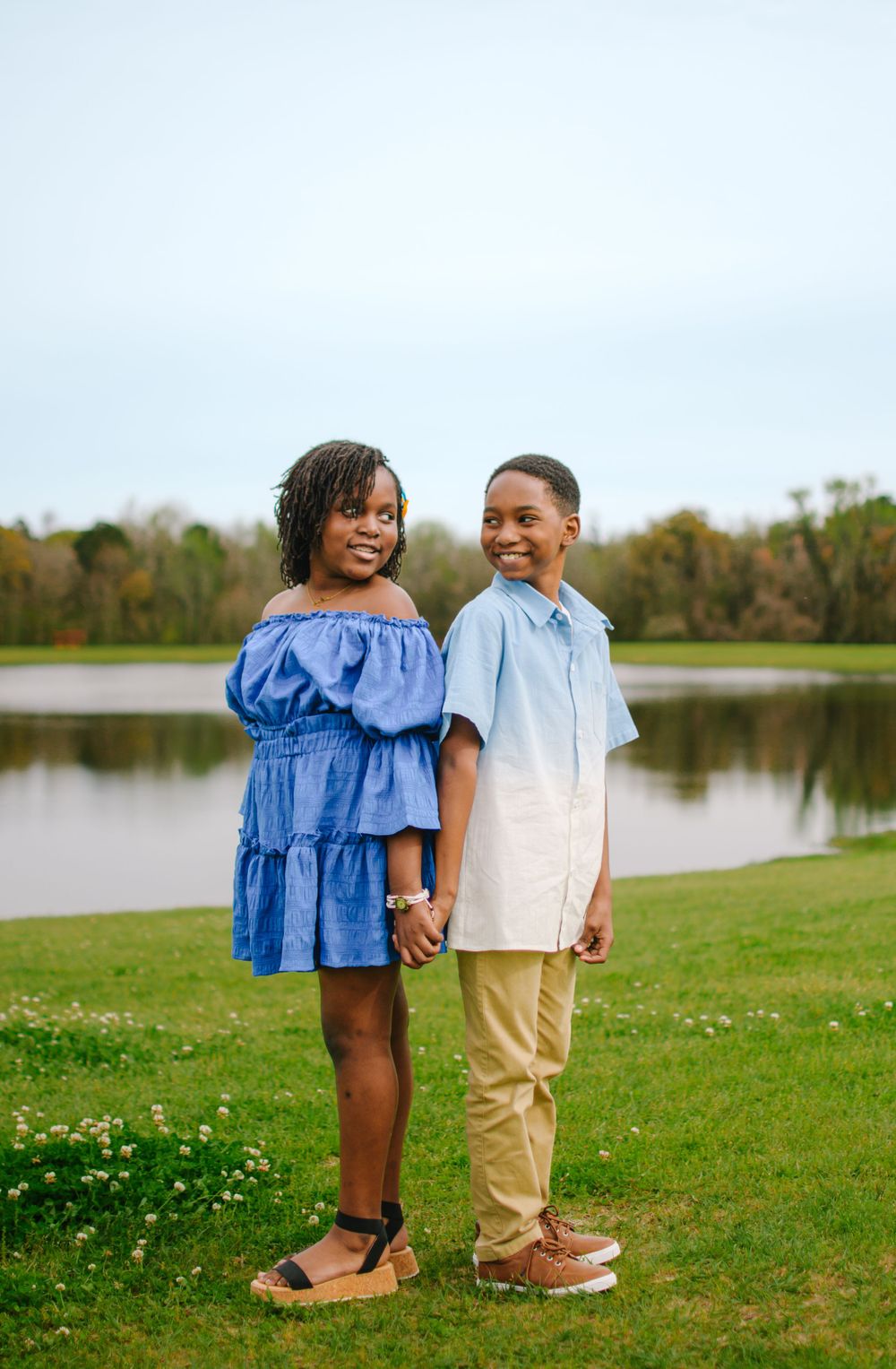 Two siblings pose together by a peaceful lake on a sunny day wearing coordinated blue and beige outfits.