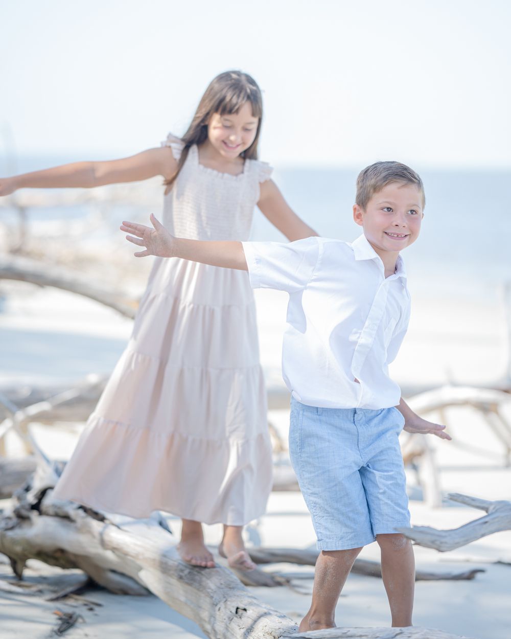 Two children playing and balancing on driftwood at a sunny beach in light summer clothing.