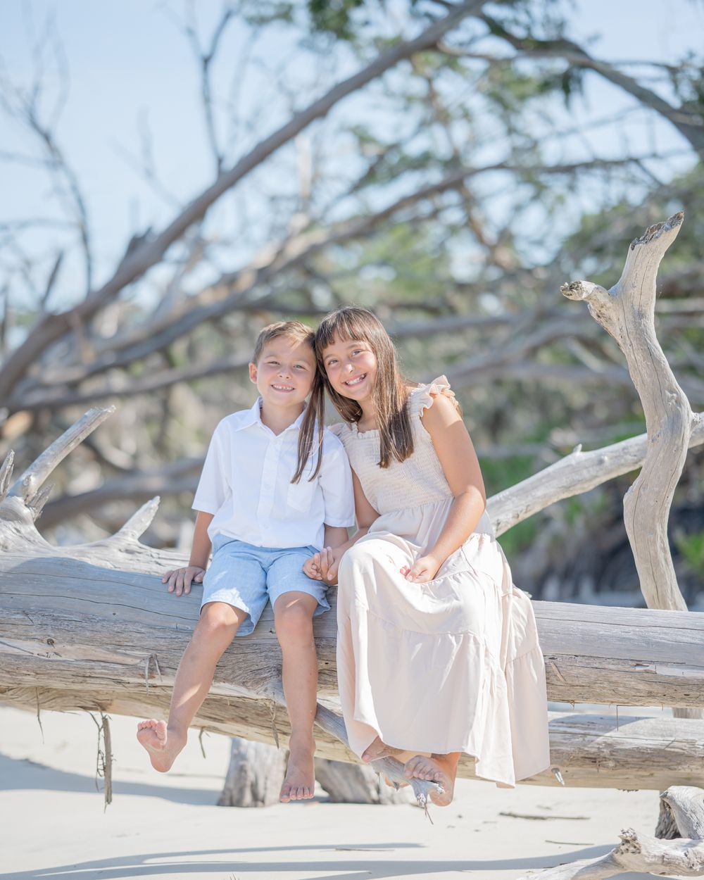 A pair sits together on a weathered driftwood log at a sunny beach, wearing white and beige casual outfits.