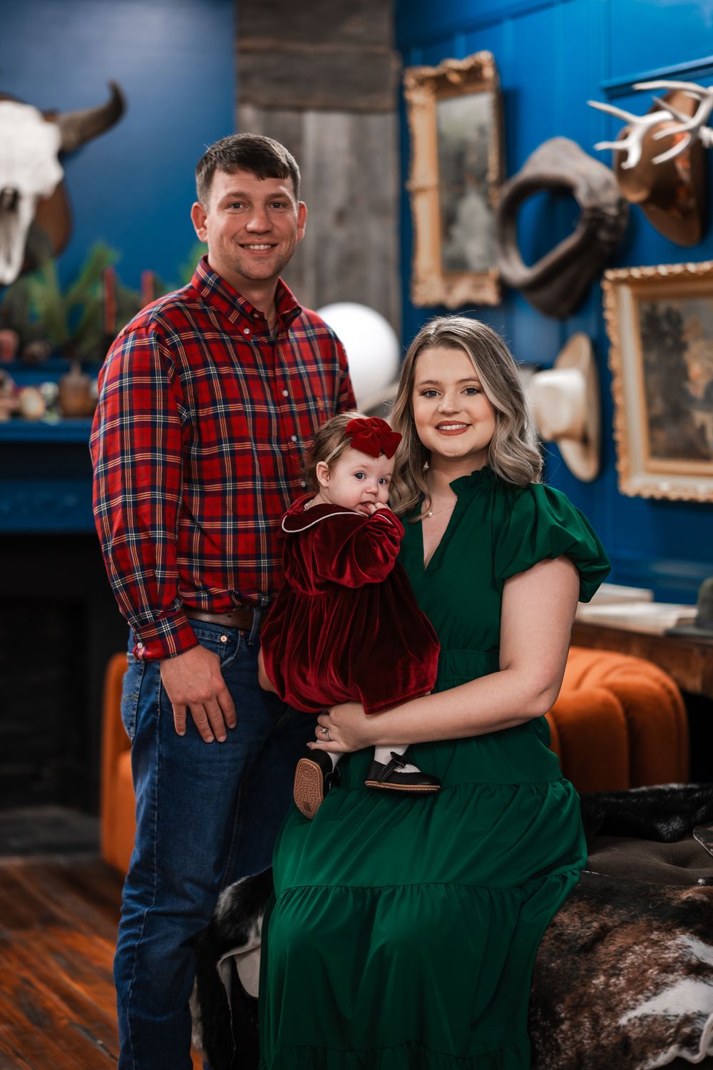 Family of three poses in a rustic setting against a blue wall with mounted decor, wearing red and green attire.