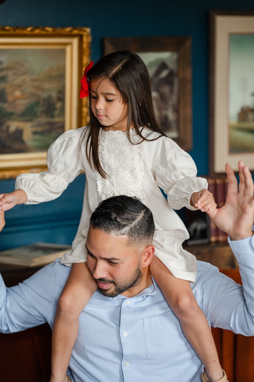 A child in a white Victorian-style dress sits on someone's shoulders in a room with framed artwork on blue walls.