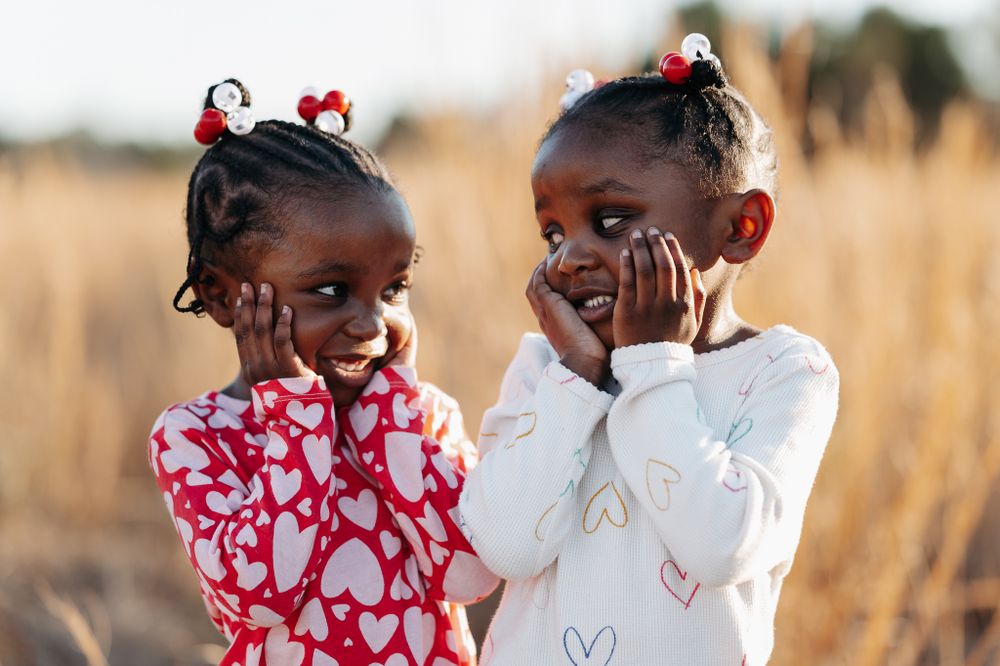 Two young children wearing red and white outfits stand together giggling and playing in a golden field at sunset.