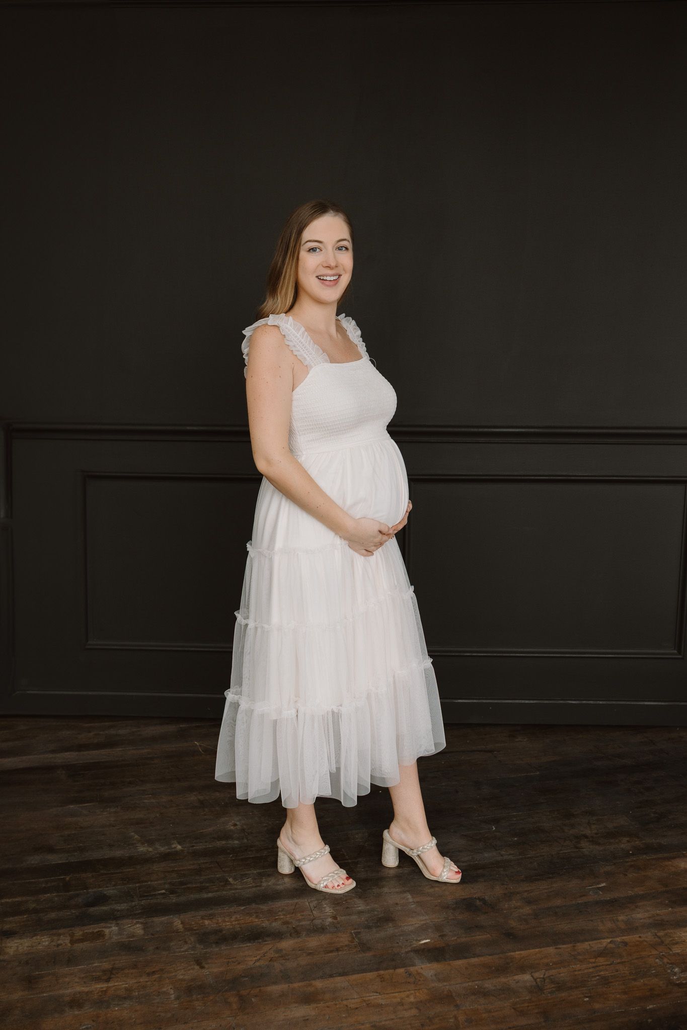 A pregnant woman is standing in a studio in front of a black wall in a white ankle-length dress touching her belly and smiling.