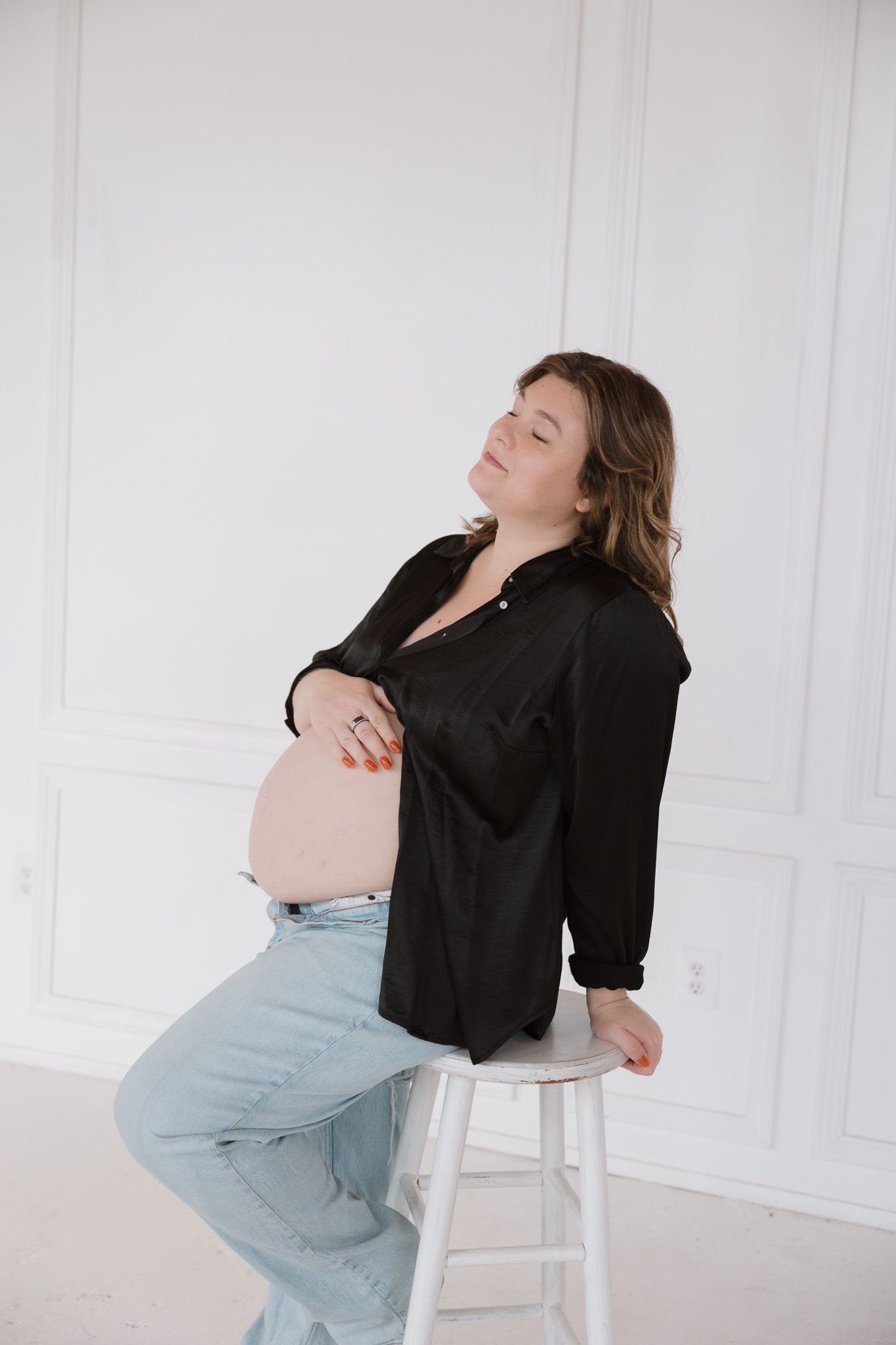 A pregnant woman in jeans and an open black blouse is leaning against a white barstool touching her belly and smiling.