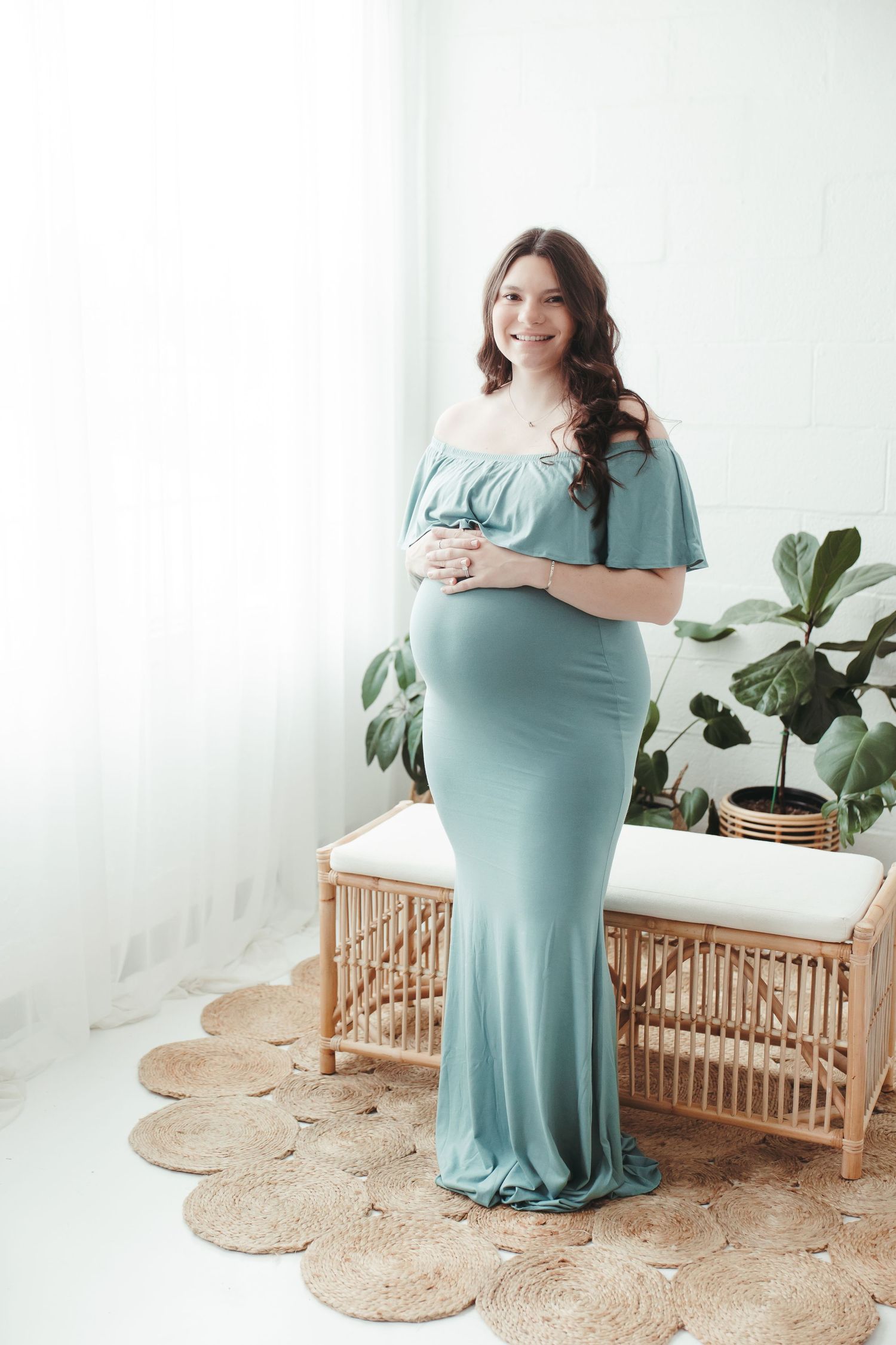 A pregnant woman is standing inside a studio in front of a window in a teal floor-length dress touching her belly and smiling.