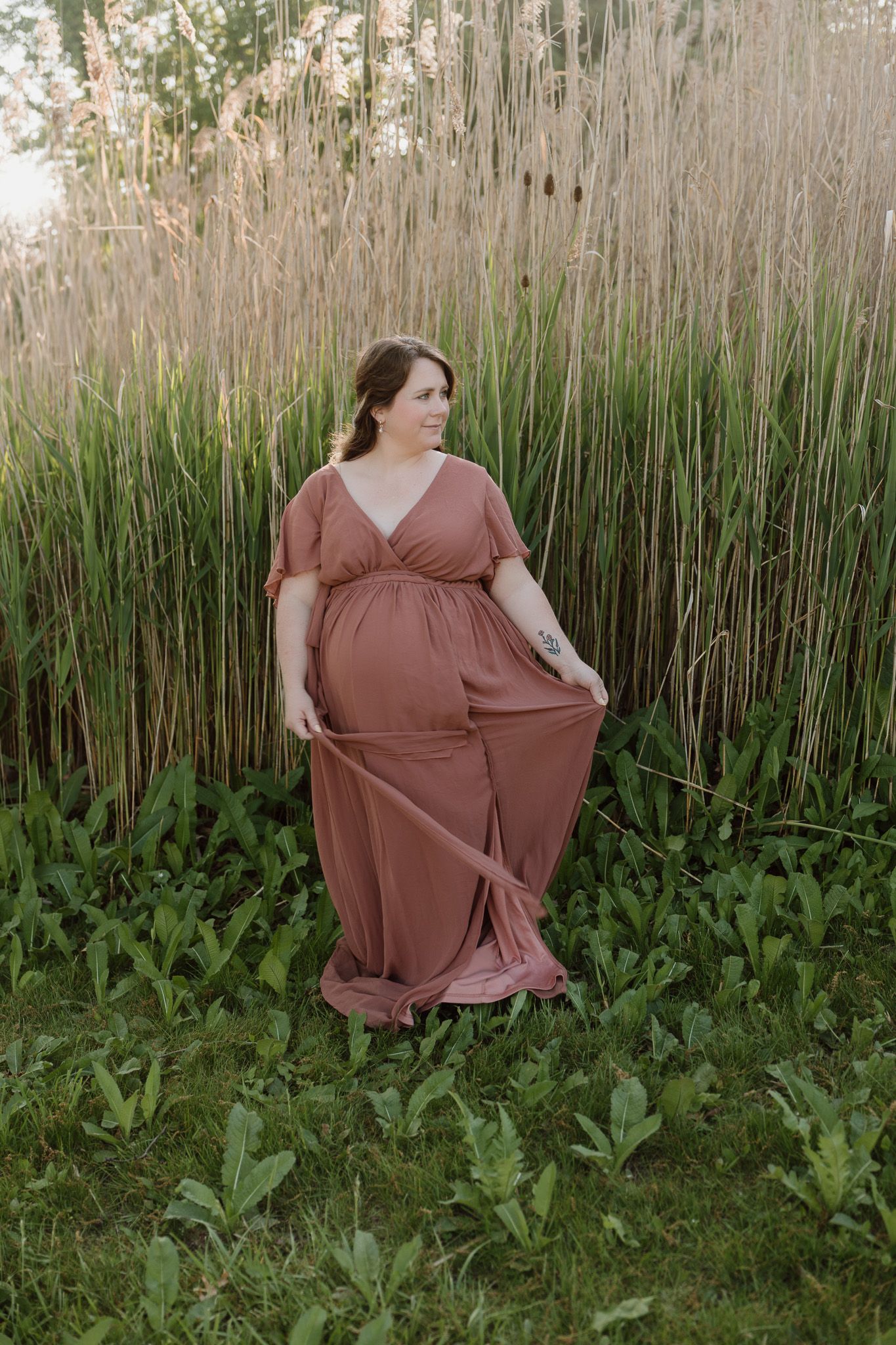 A pregnant woman is standing outside in front of tall grasses in a pink floor-length dress swaying her dress and smiling.