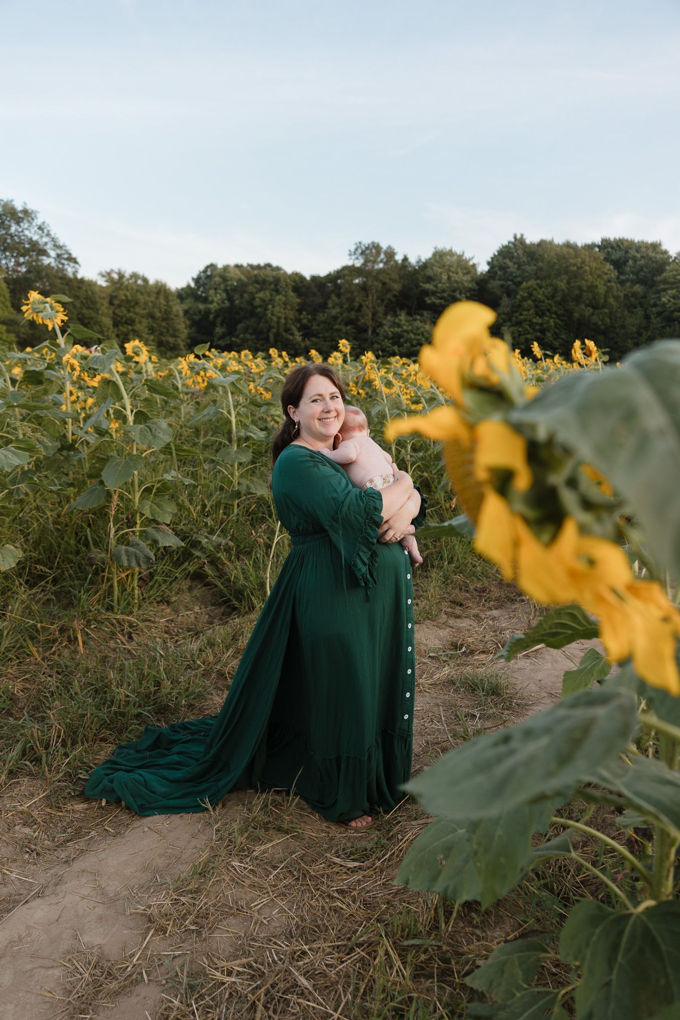 A pregnant woman is standing outside in a sunflower field holding a baby on her shoulder and smiling