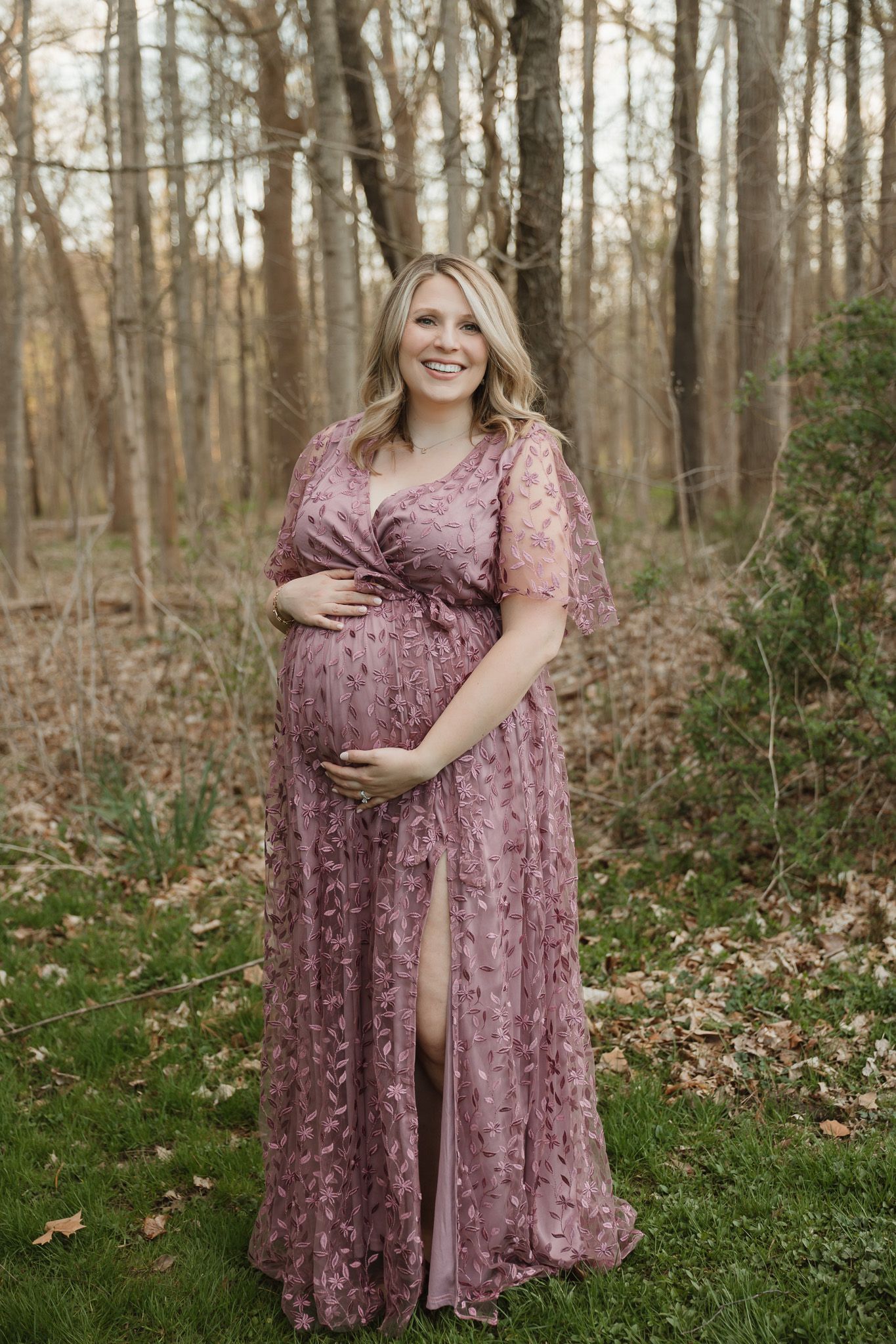 A pregnant woman is standing outside in front of a tree line in a purple floor-length dress touching her belly and smiling.