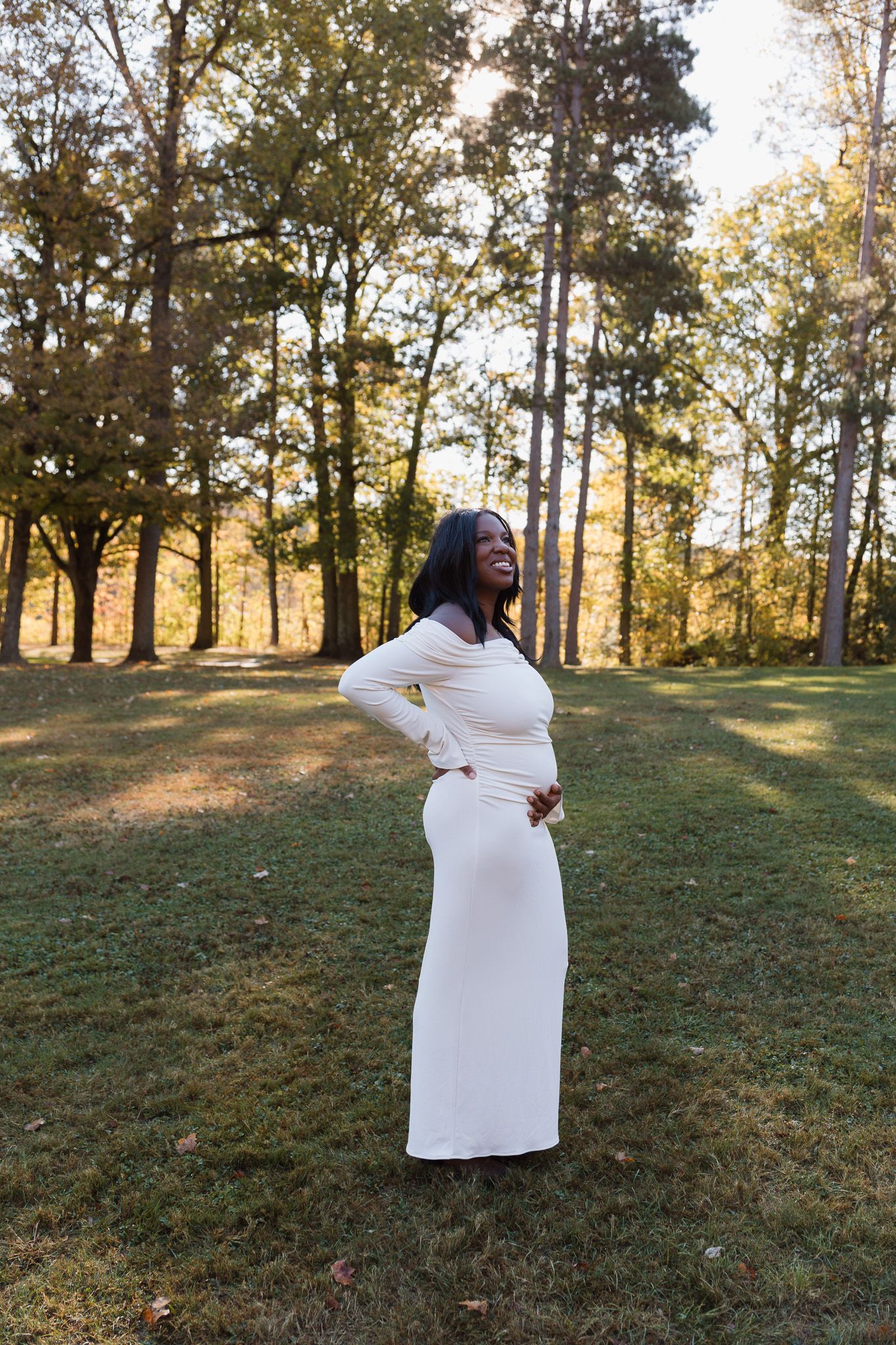 A pregnant woman is standing outside in front of a tree line in a white floor-length dress touching her belly and smiling.
