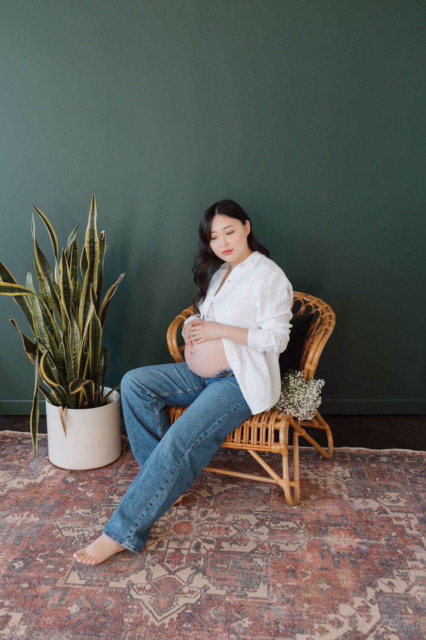 A pregnant woman in jeans and an open white button down blouse is seated in a chair holding her belly