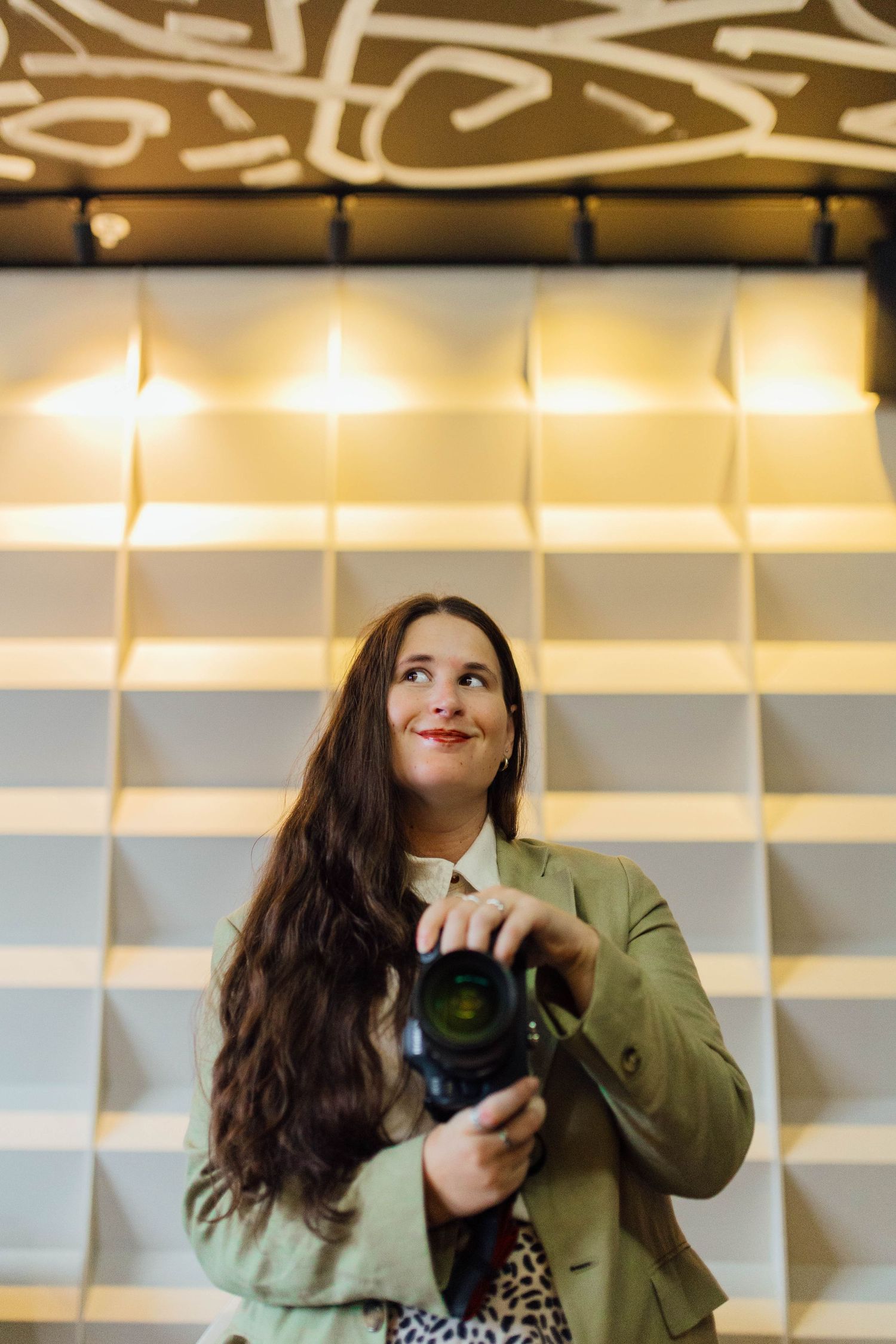 Woman in green jacket holding DSLR camera, looking up thoughtfully against a modern geometric lit wall backdrop.