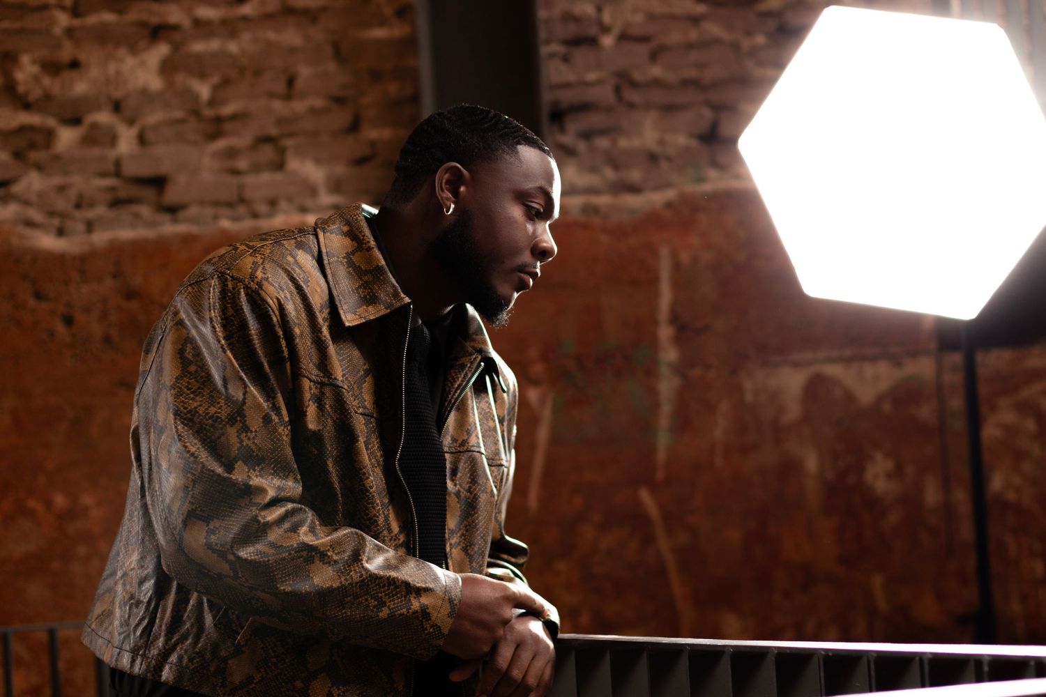 A man in a gold jacket leans on a railing under a studio light against a rustic brick wall.