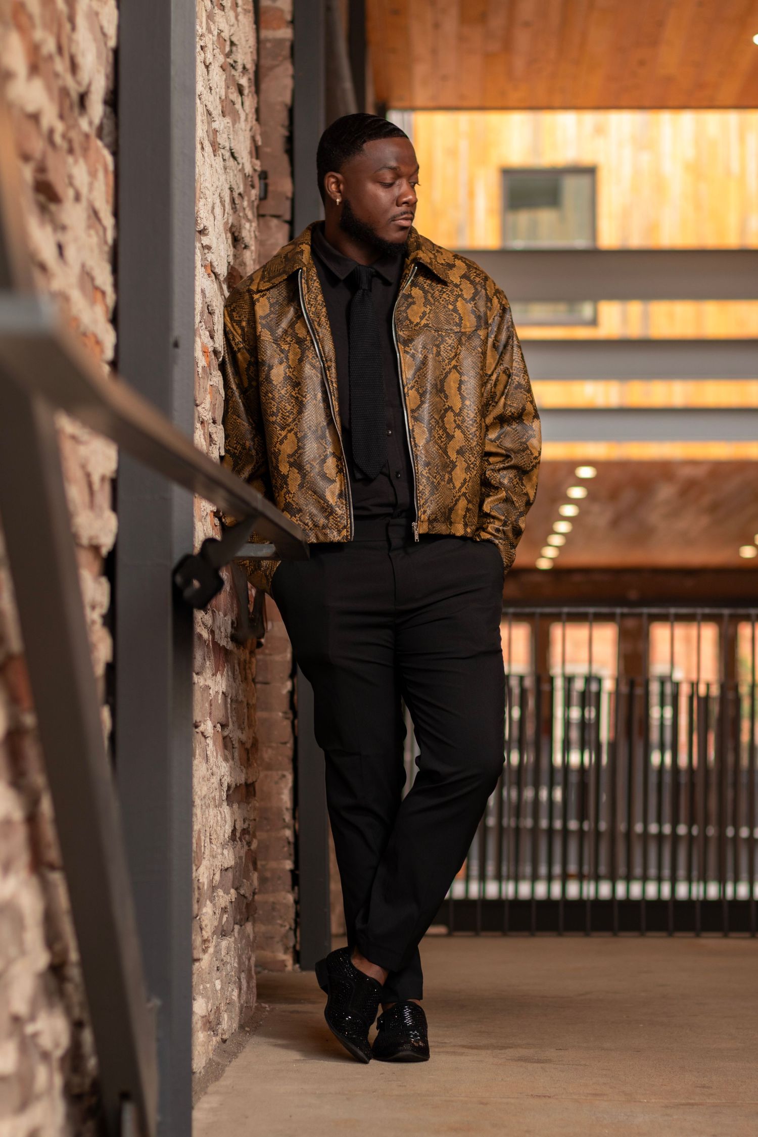 Man in snakeskin jacket and black pants leaning against brick wall in modern industrial setting.