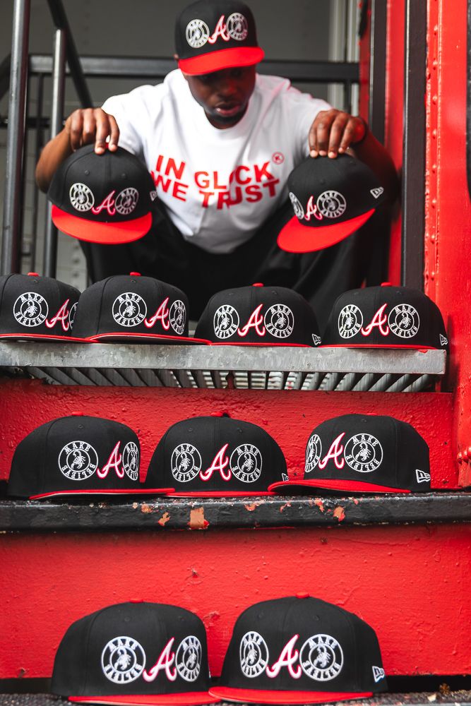 Man in white tee displays black and red New Era Atlanta Braves snapback hats on red metal stairs.