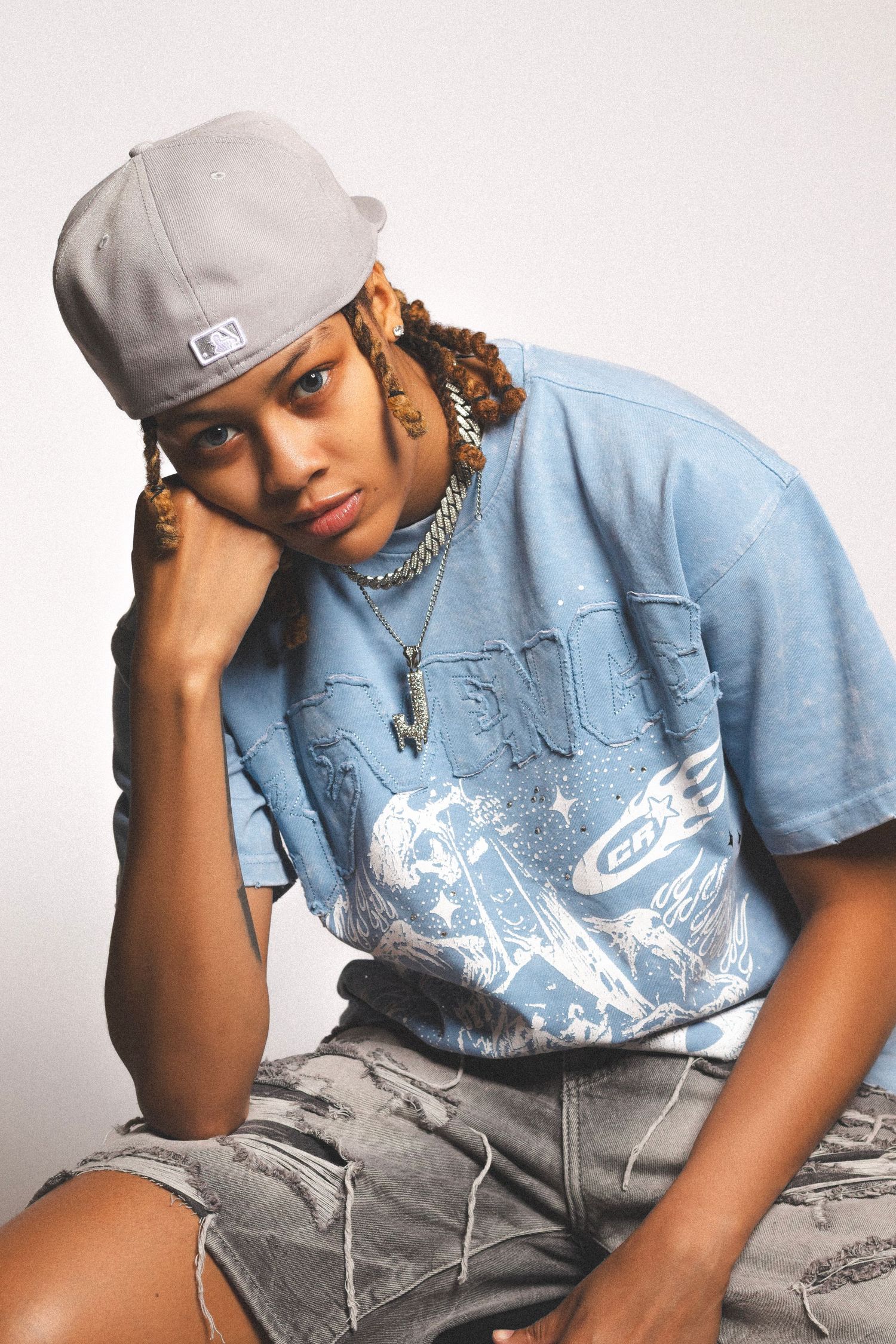 Young person in gray snapback, blue graphic tee, and denim shorts posing confidently in a studio setting.