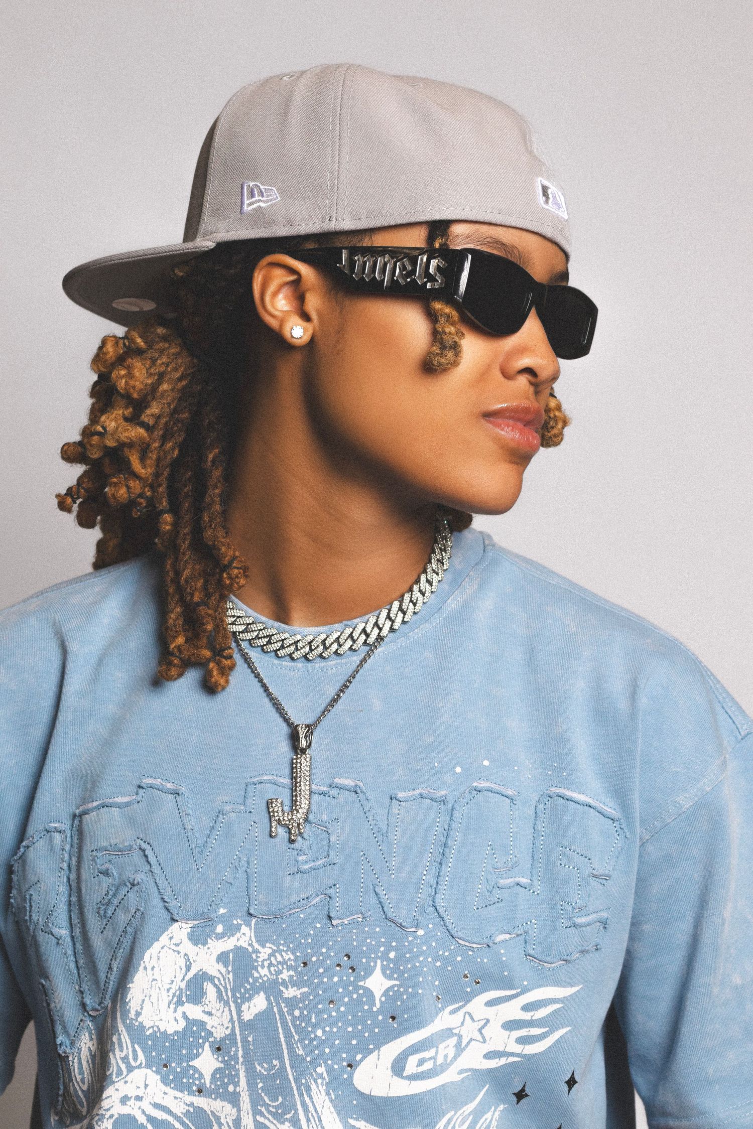 Young person wearing gray New Era cap, sunglasses, chain necklace, and blue graphic sweatshirt with locs hairstyle.