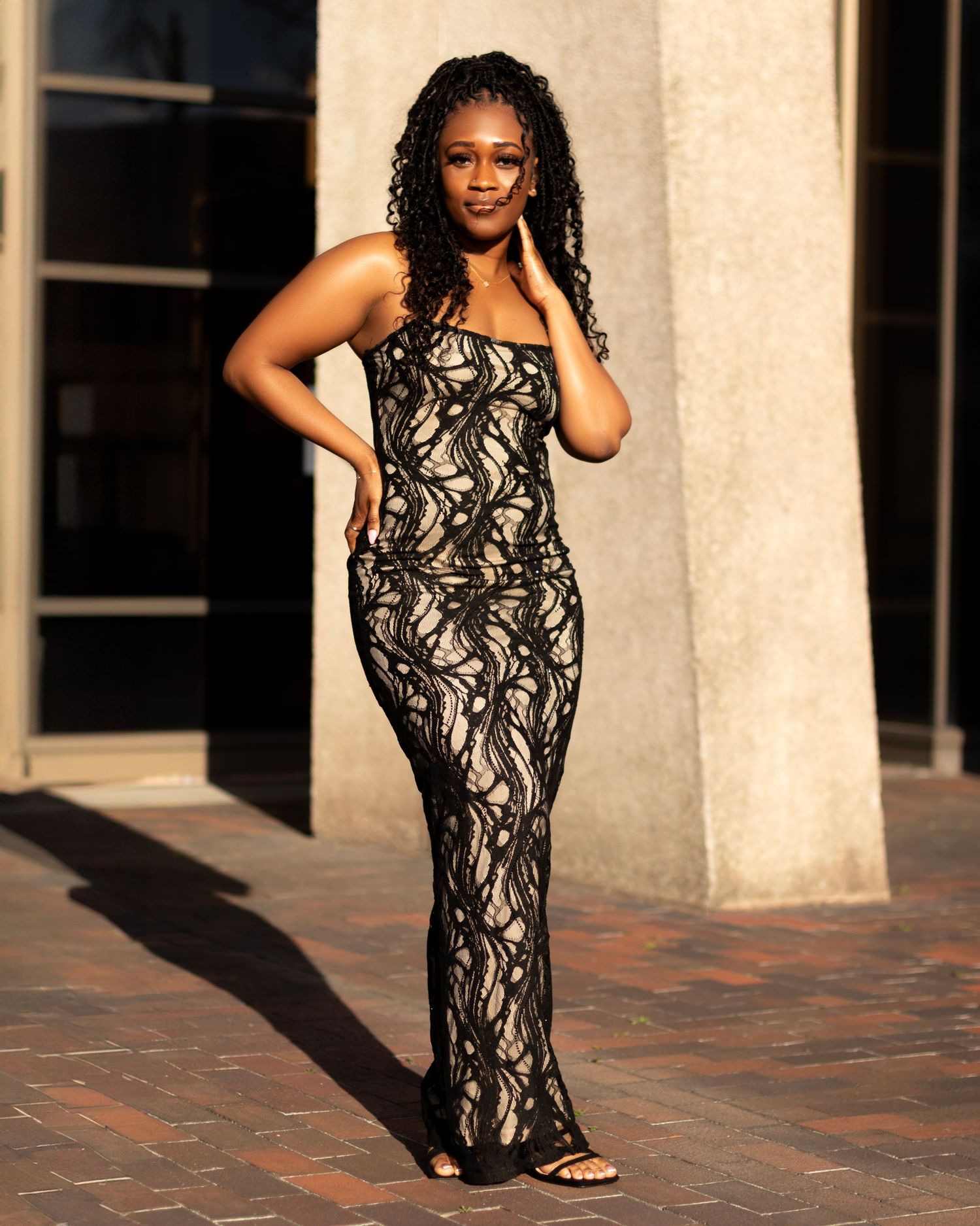 Woman in black lace floor-length gown posing outdoors in golden sunlight against a concrete building.