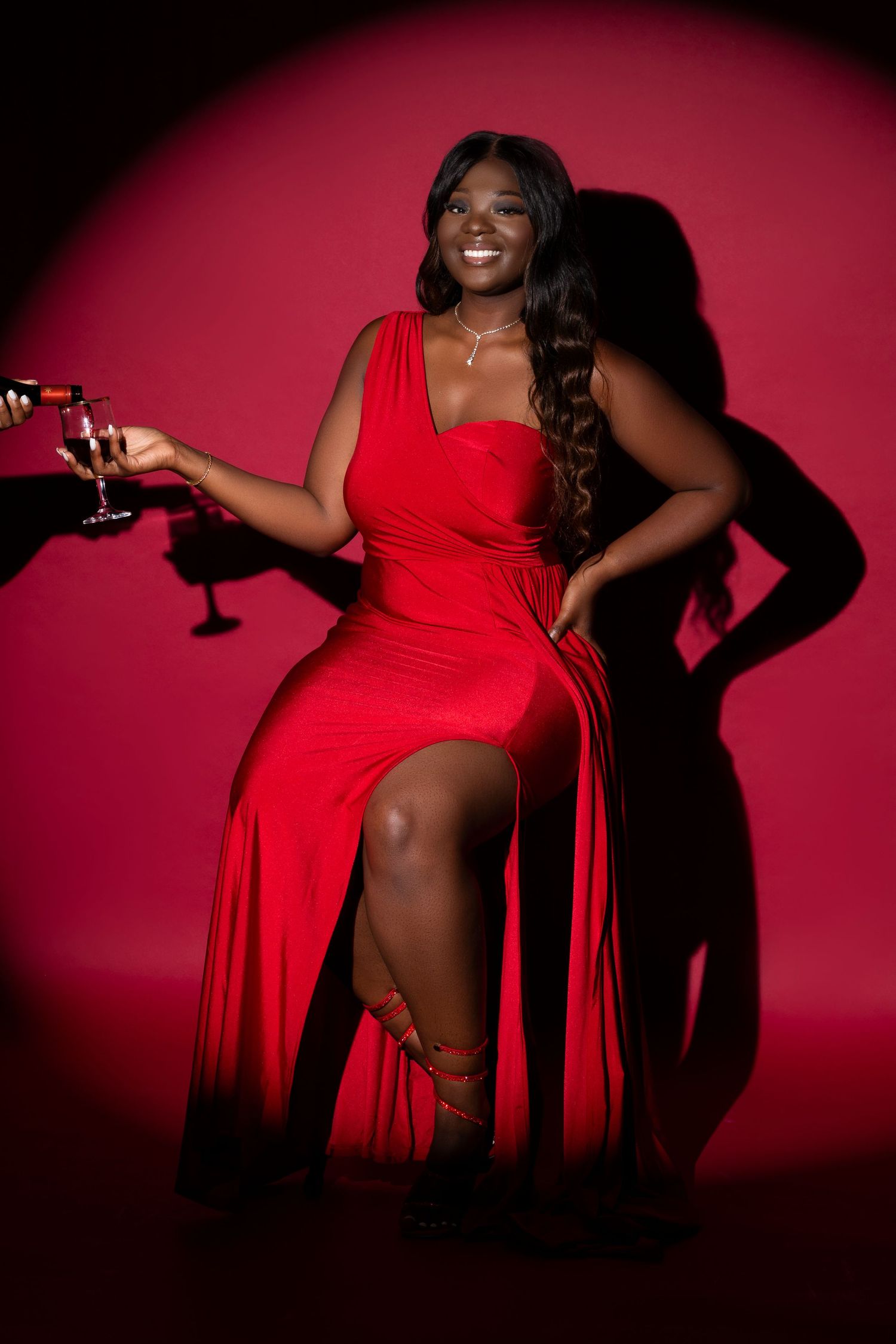 Elegant pose in red evening gown holding champagne glass against red backdrop.