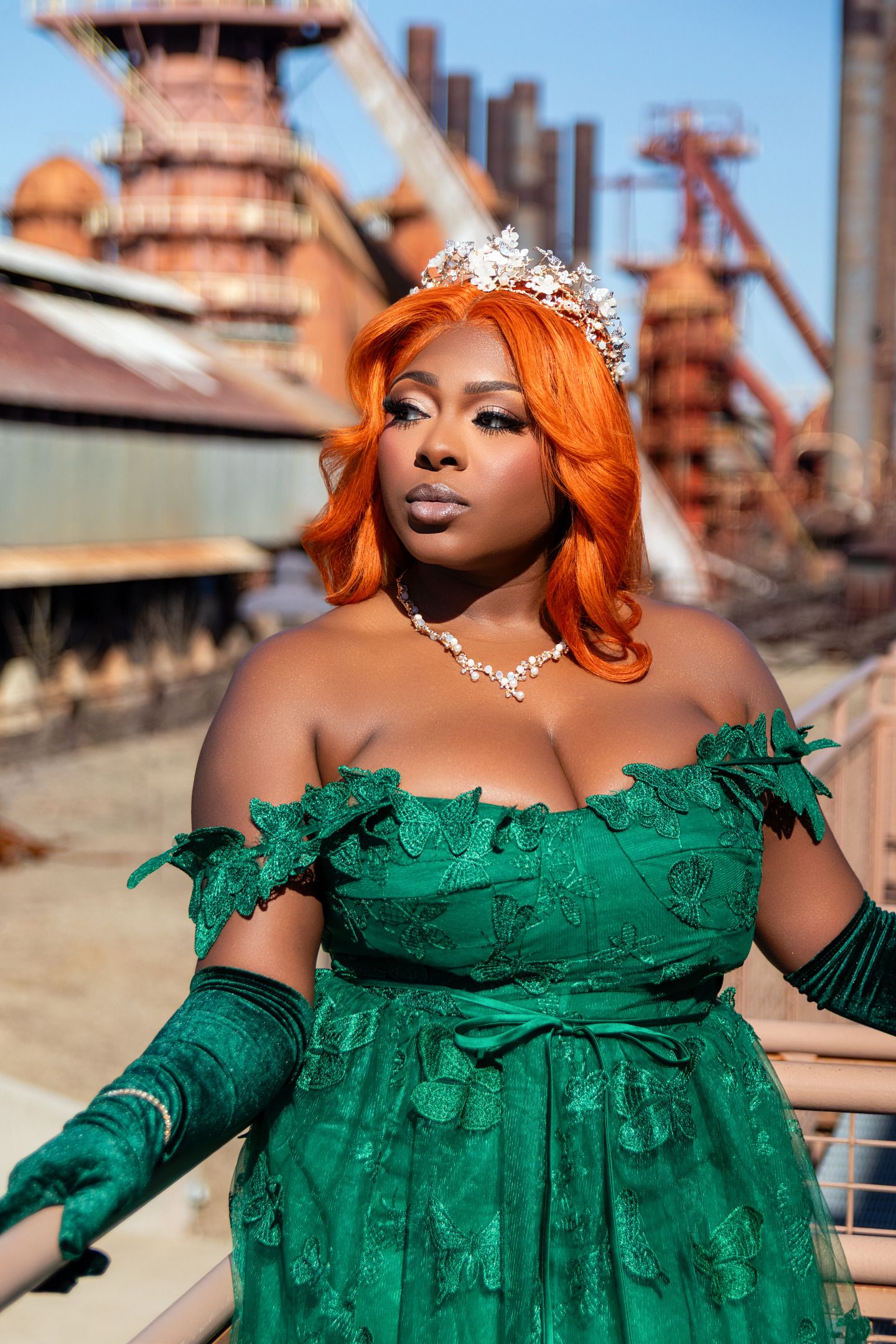 Woman in green off-shoulder gown, orange hair, crystal crown and necklace, posing outdoors near industrial structure.