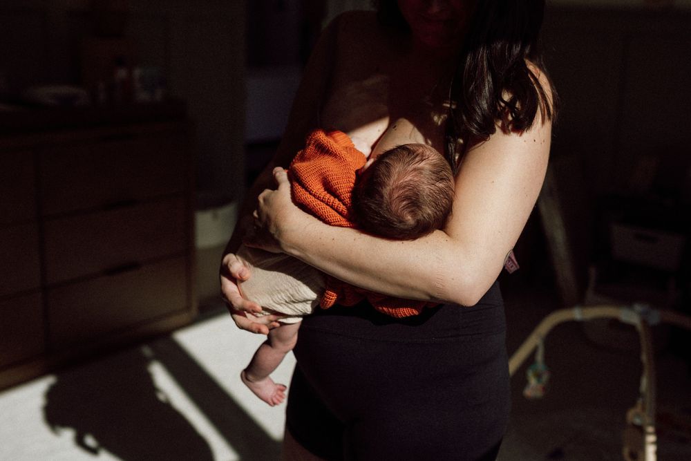 A parent in a dark room holds a newborn baby wearing an orange sweater while sunlight streams through the window in suffolk, virginia