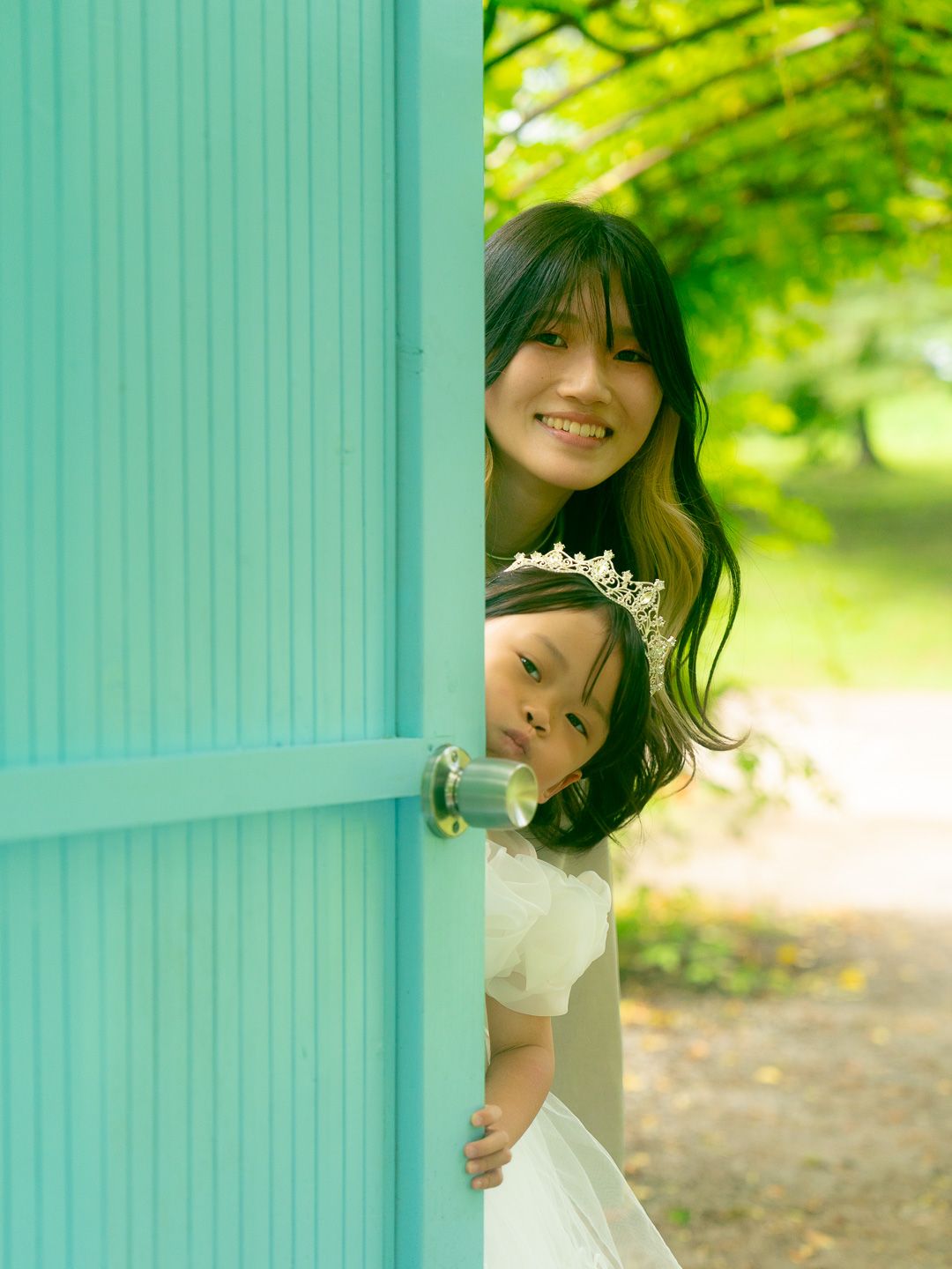 A smiling woman and young girl wearing a tiara peek playfully around a turquoise door in a lush green park.