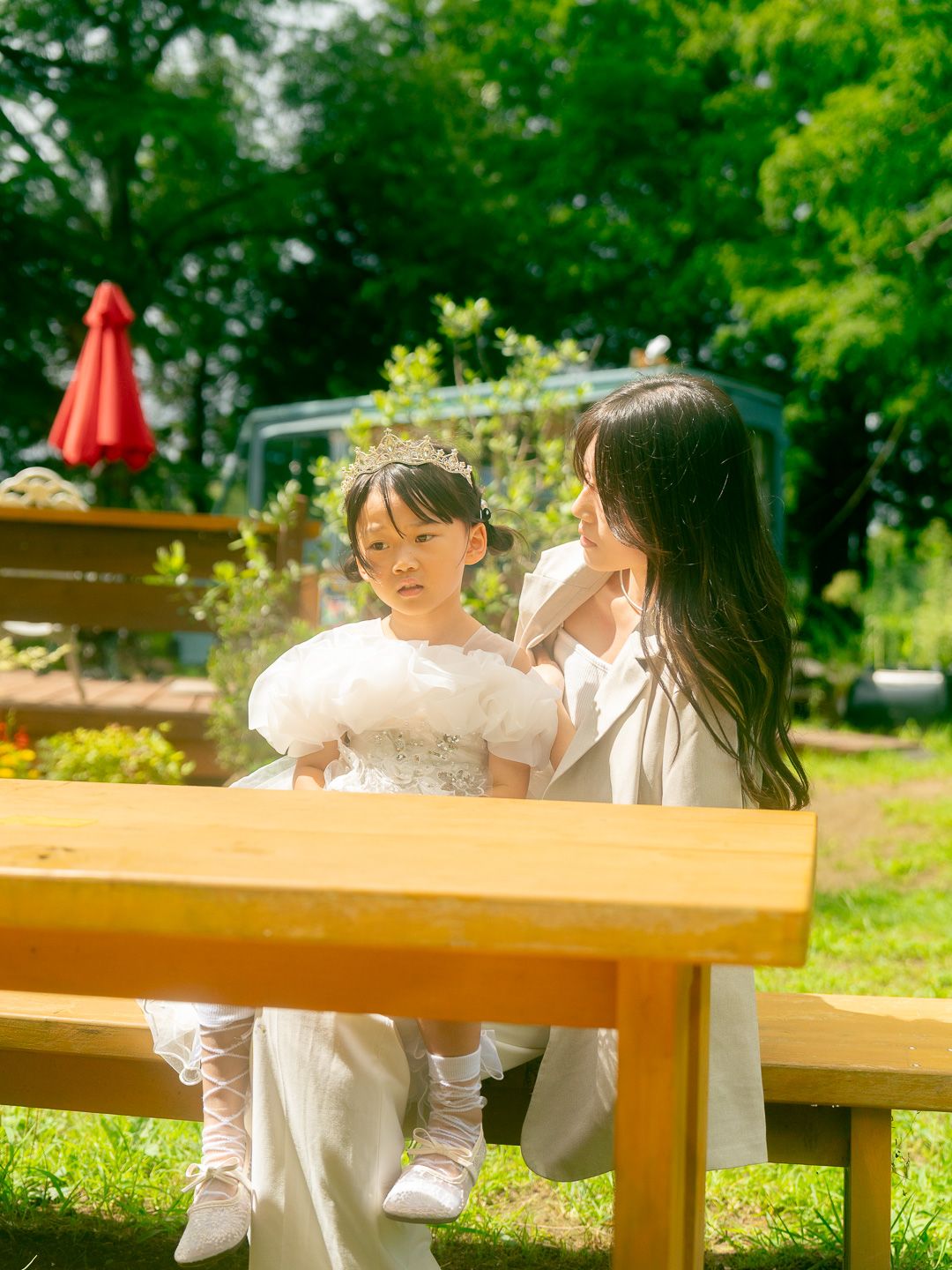A woman in white adjusts a young girl's dress on a sunny park bench surrounded by lush greenery.