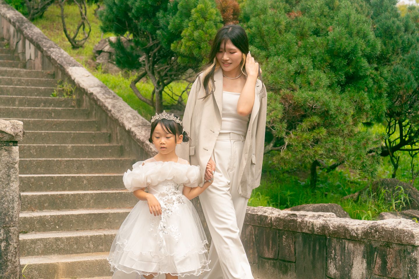 A woman and young girl in white dresses walk down stone steps surrounded by lush green trees.