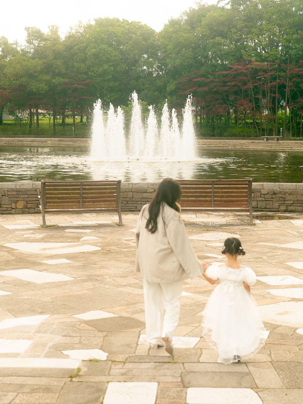 A woman and young girl in white walk hand-in-hand toward a fountain in a serene park.