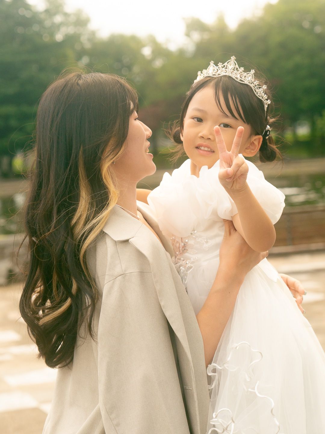 A woman holds a young girl wearing a white dress and tiara, who flashes a peace sign, outdoors.