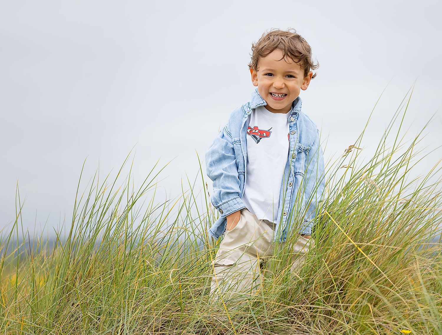 Young child in blue jacket smiling among beach grass.