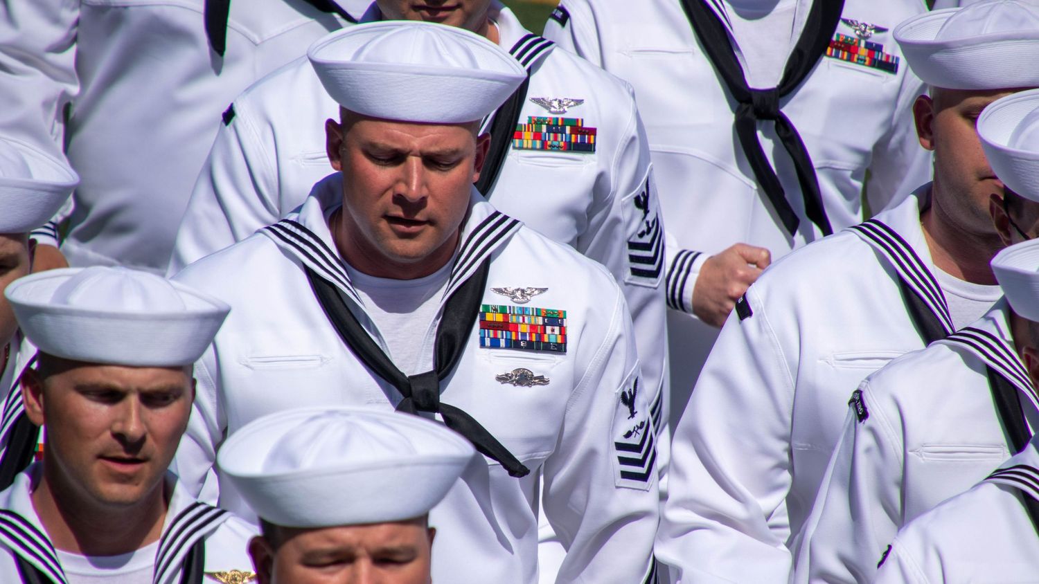 Close-up military event photography of a formation of U.S. Navy Sailors marching in step wearing Service Dress White uniforms.