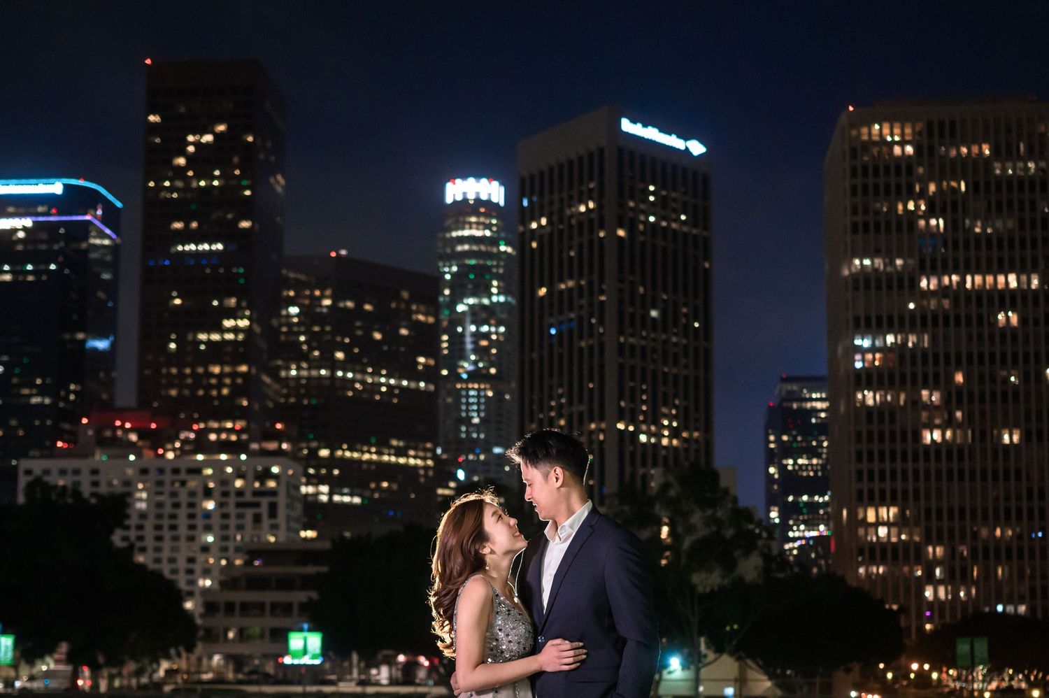 A couple embraces against a stunning downtown city skyline lit up at night.